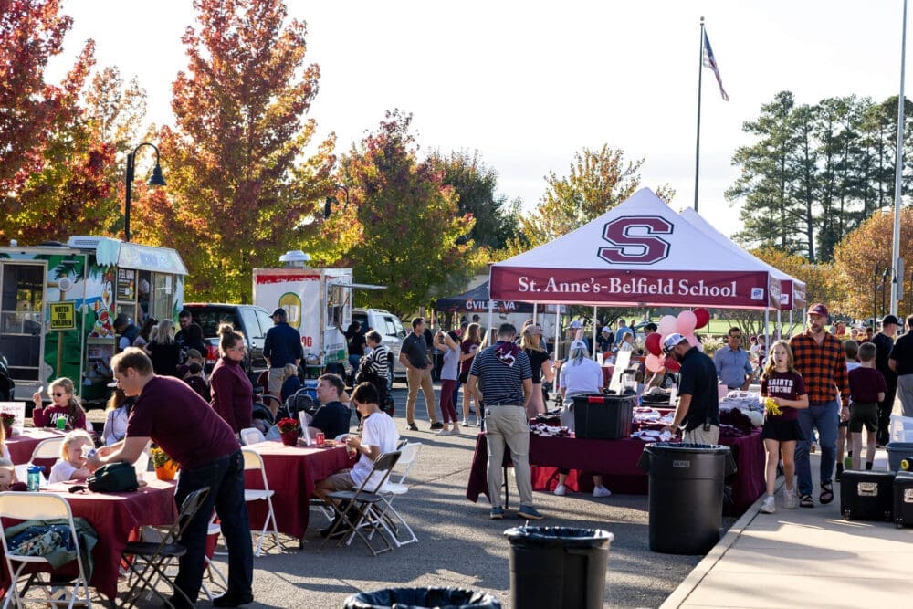 People gather outdoors at a school event with tables, food trucks, and a St. Anne’s-Belfield School tent. Trees with autumn leaves are in the background, and attendees are socializing and eating.