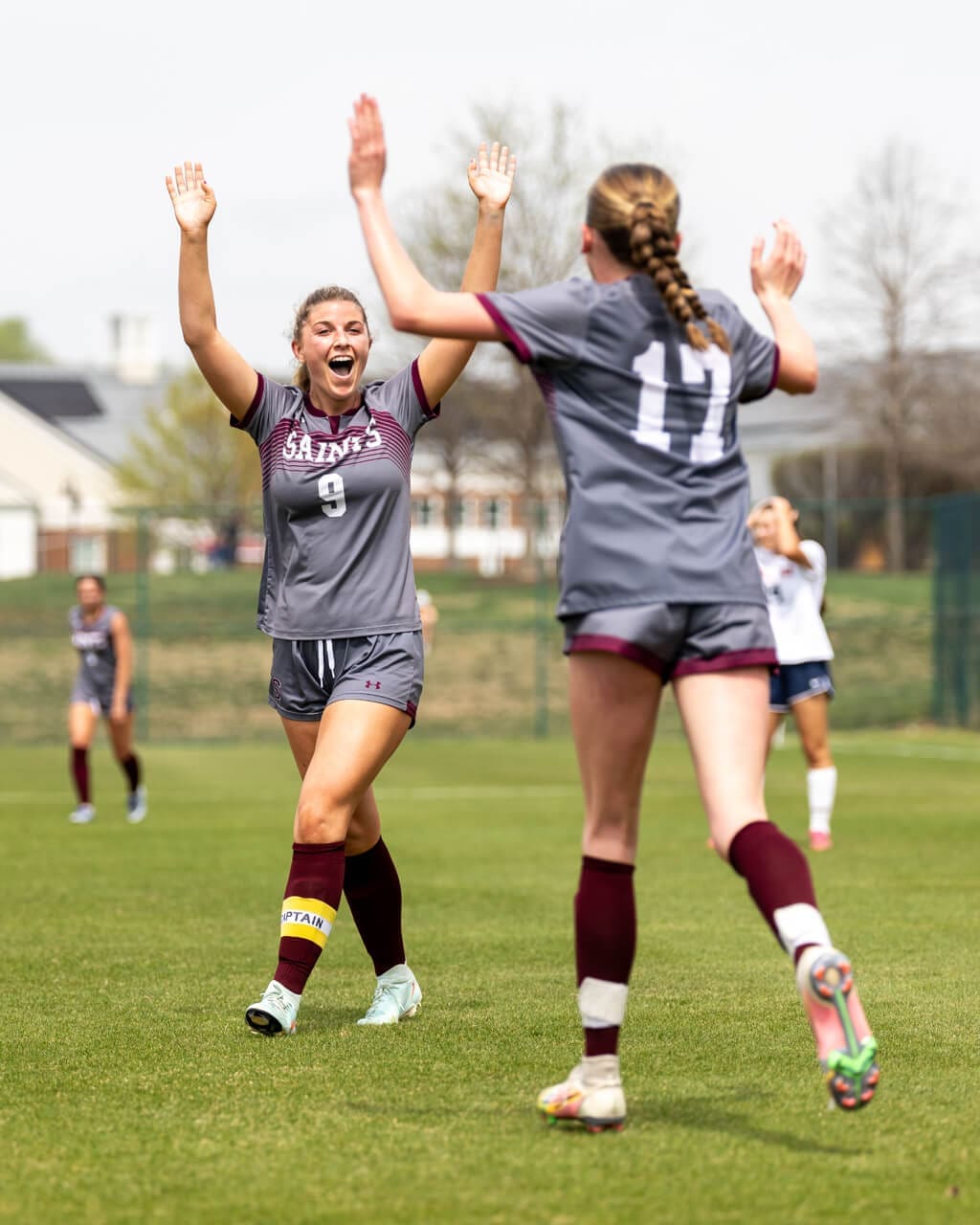 Two female soccer players in gray school uniforms celebrate on a grassy field, facing each other with arms raised for a high-five. Other players and trees are visible in the blurred background.