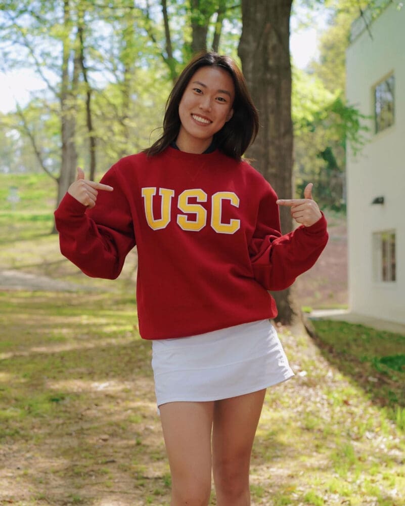 Cici outdoors, smiling and pointing at her red sweatshirt with large yellow USC letters. She is wearing a white skirt and is surrounded by trees and greenery.