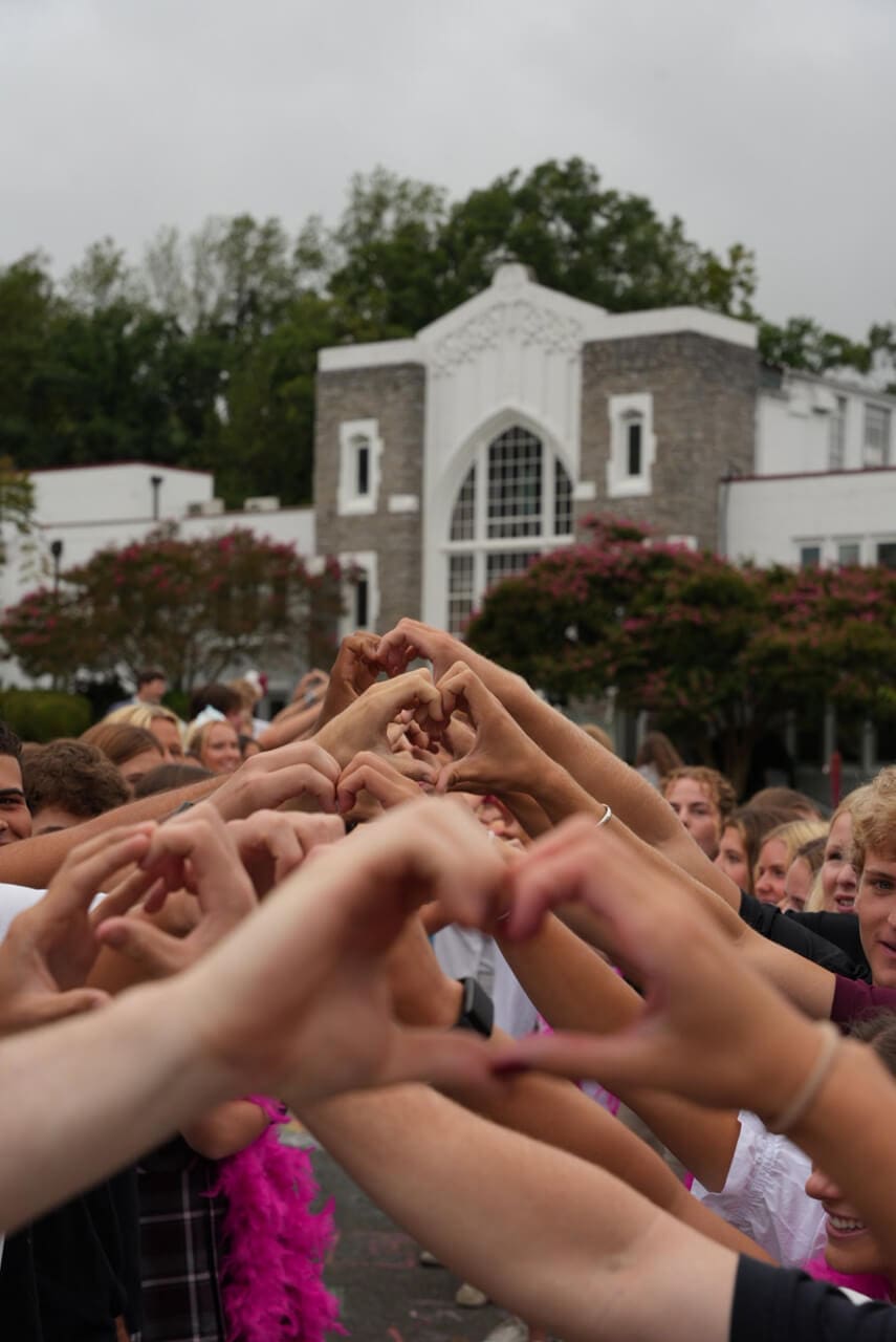 A group of people stand outdoors at a school, forming heart shapes with their hands to create a tunnel. A large building with arched windows and lush greenery is visible in the background under a cloudy sky.
