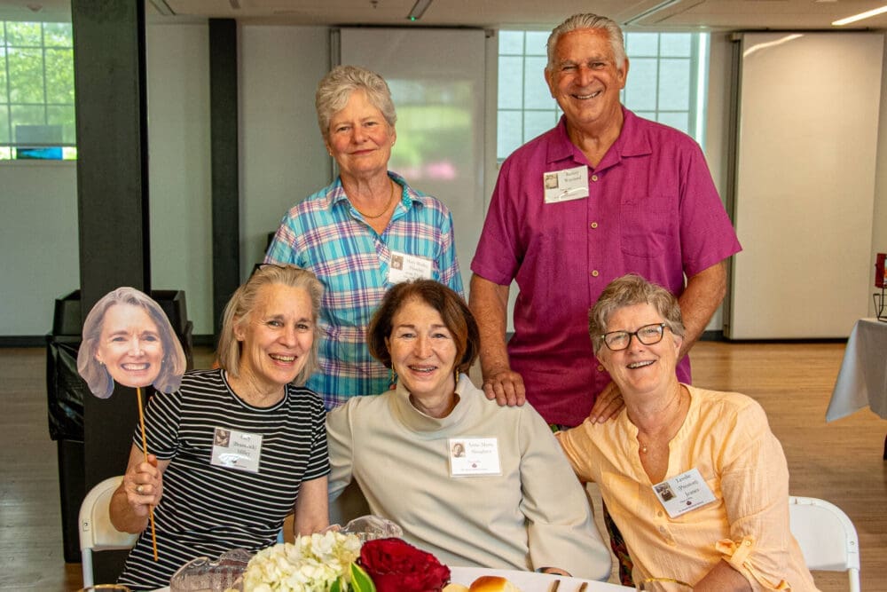 Five smiling older adults pose together at a table indoors. Four women are seated, one holding a cutout of a womans face, while a man stands behind them. Name tags are visible on their shirts.