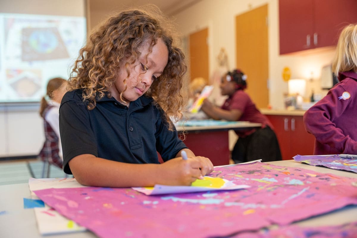 A young child with curly hair draws on colorful paper at a classroom table, with other children working in the background.