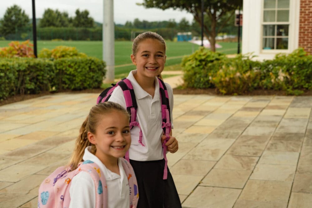 Two smiling school-age girls wearing white polo shirts and backpacks stand outside on a paved walkway near greenery and a brick building, likely on their way to school.