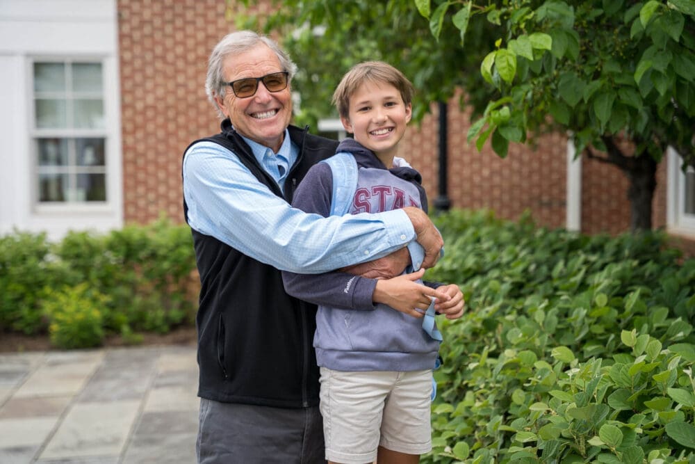 An older man and a smiling child stand outside near green bushes. The man, wearing glasses and a vest, hugs the child, who holds a basketball and wears a hoodie and shorts. Both look happy and are facing the camera.