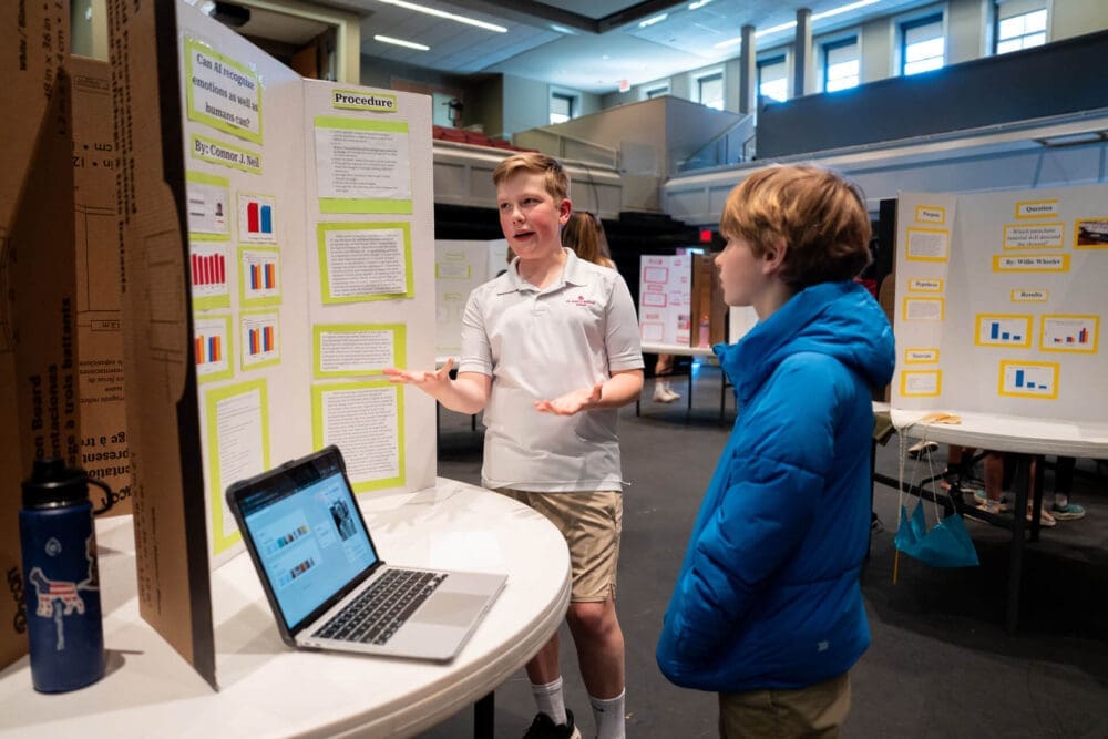 A boy presents his science project to another student at a school science fair, standing by a display board with graphs and text. A laptop and a water bottle are on the table. Other project displays are visible in the background.