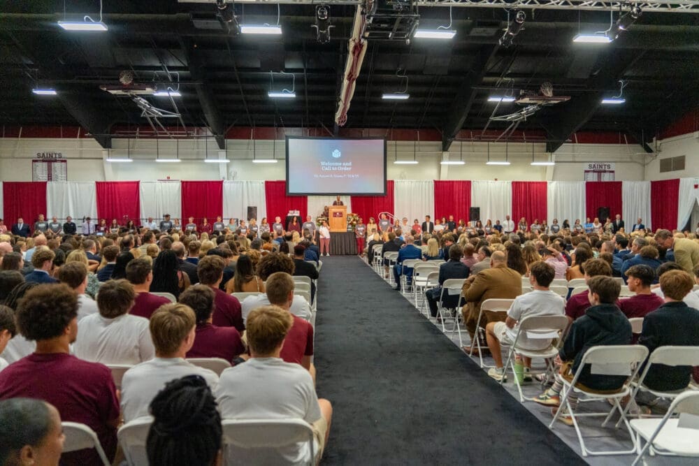 A large group of people sit in rows of chairs facing a stage in a gymnasium, where a speaker stands at a podium. A screen above the stage displays text. The audience appears to be students and adults.