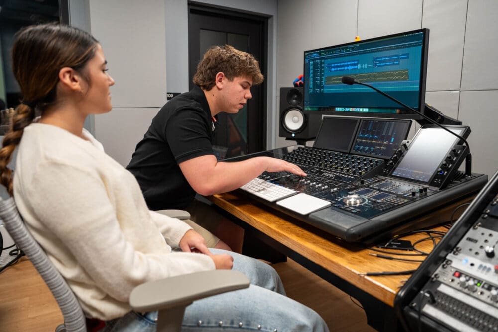 Two people sit in a recording studio; one operates a digital audio mixing console while the other observes. A large monitor displays audio tracks and editing software in a modern, professional school setting.