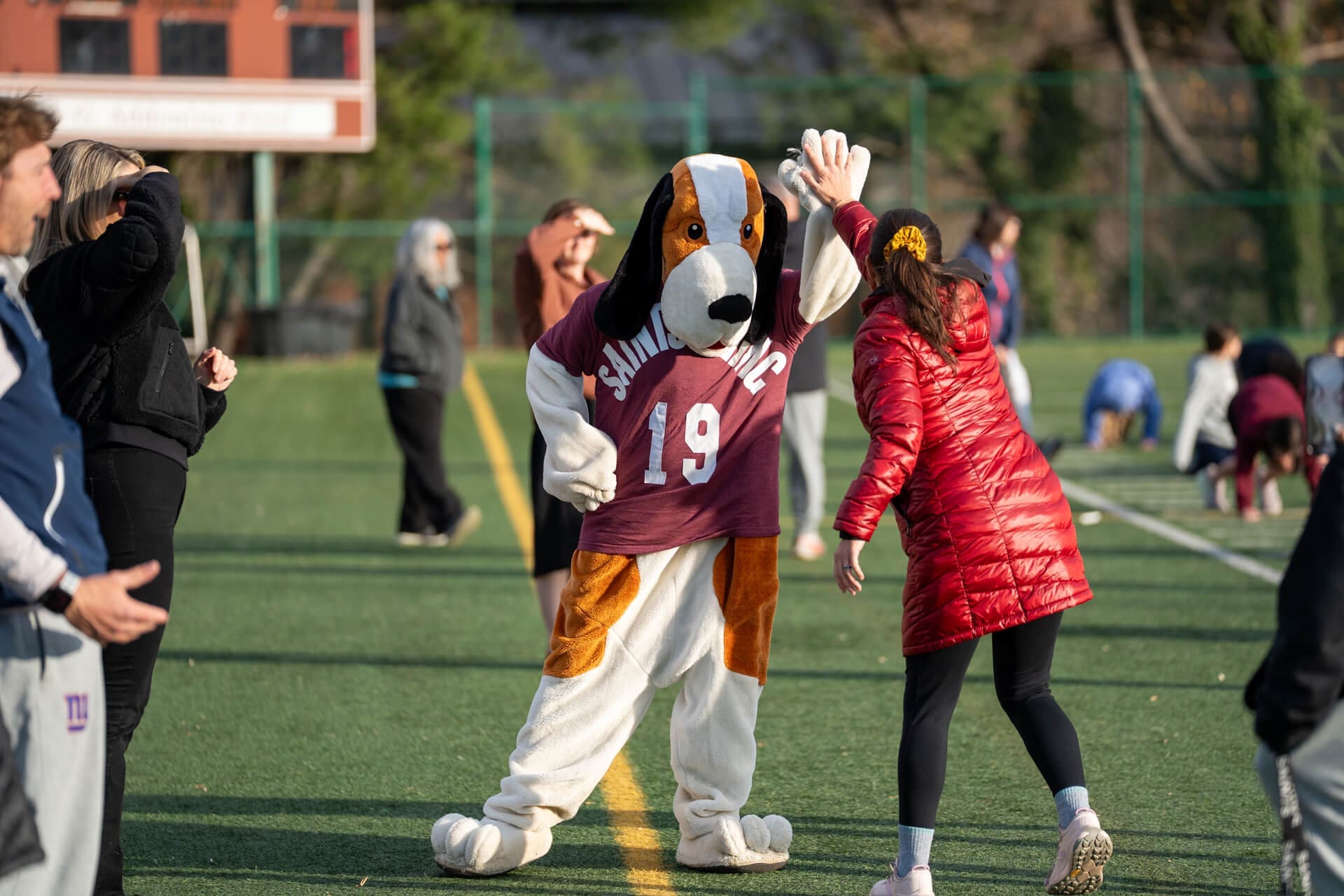 A person in a dog mascot costume wearing a maroon sports jersey high-fives a woman in a red jacket on a sports field, with other people and children in the background.