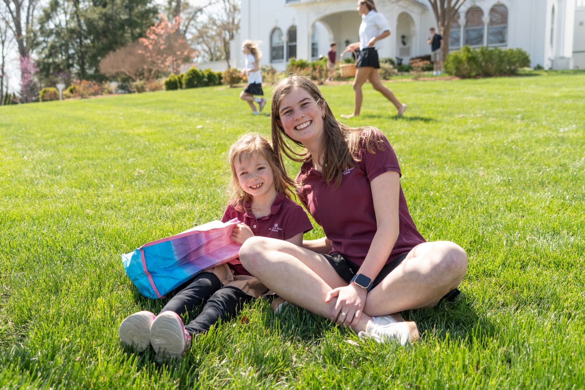 A young woman and a smiling little girl sit on green grass, both wearing maroon shirts. The girl holds a colorful bag. Other people and a white building appear in the background on a sunny day.