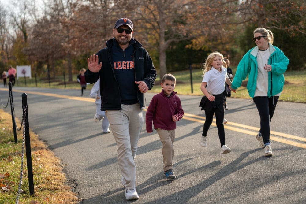 A man in a Virginia sweatshirt walks and waves on a park path with several children and a woman nearby, perhaps during a school outing. The group appears to be enjoying an outdoor activity on a sunny autumn day.