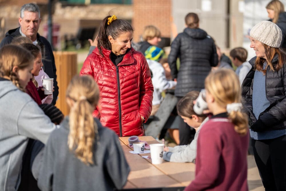A group of adults and children gather around a wooden table outdoors on a sunny day, talking and drinking from cups. The focus is on two women in bright jackets smiling and engaging with the children.