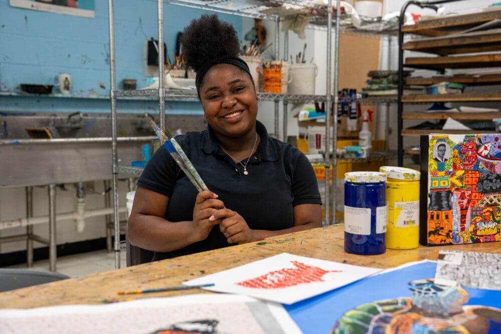 A smiling young woman sits at a table in an art studio, holding paintbrushes. Paint cans, colorful artwork, and art supplies are on the table and shelves are visible in the background.