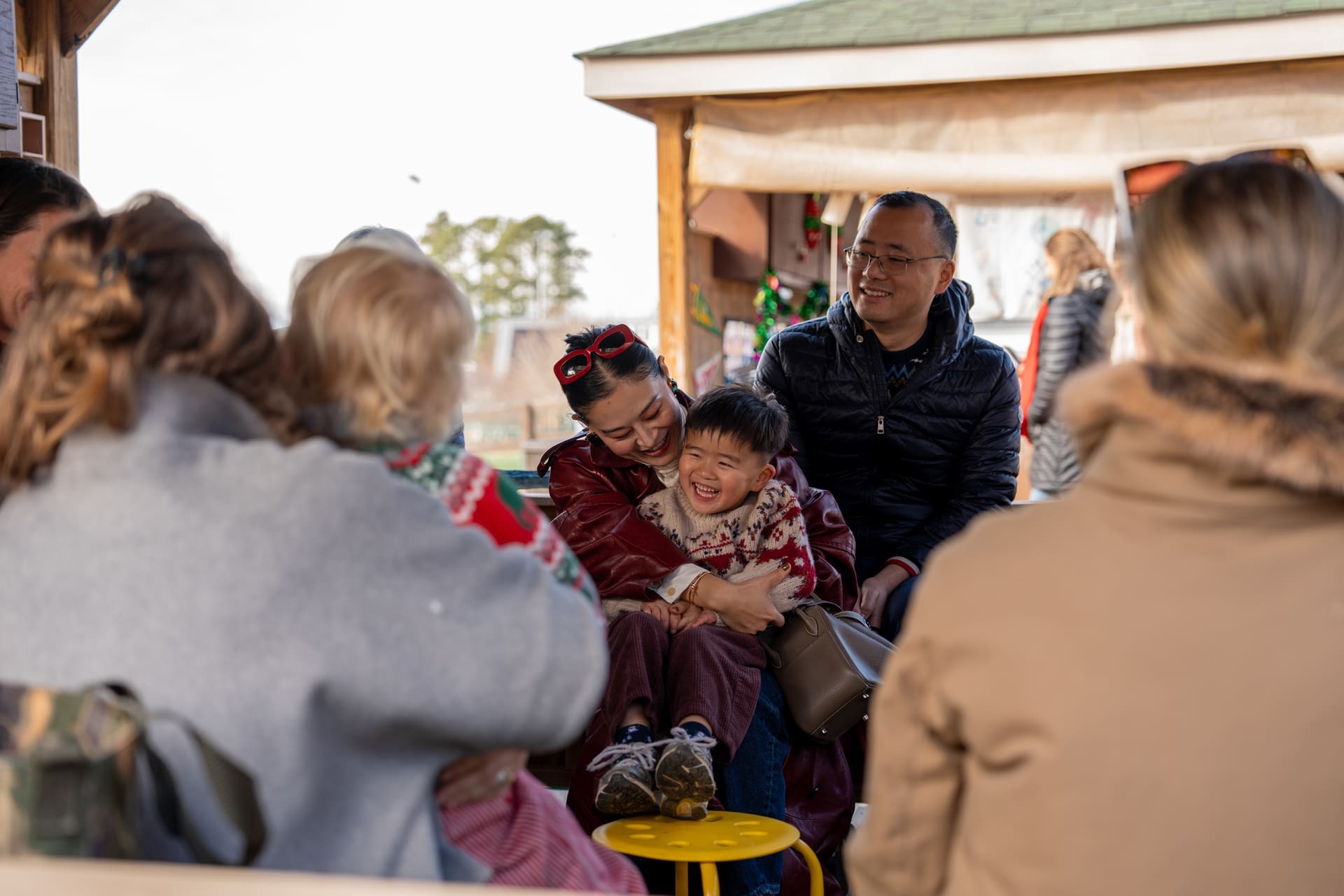 A group of people sit closely together outdoors. A woman in red sunglasses hugs a smiling young boy while others chat nearby. The setting appears festive and cheerful, possibly at a holiday event.