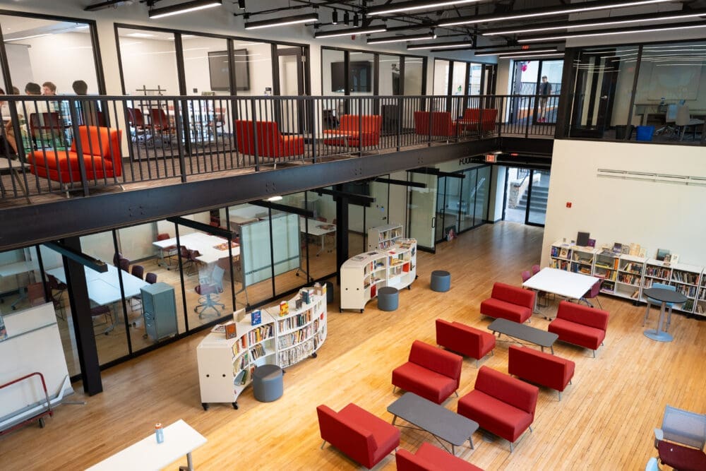A modern library interior with two levels, featuring red lounge chairs, bookshelves, tables, and large glass walls. The upper level has more seating and overlooks the spacious lower area with wooden flooring.