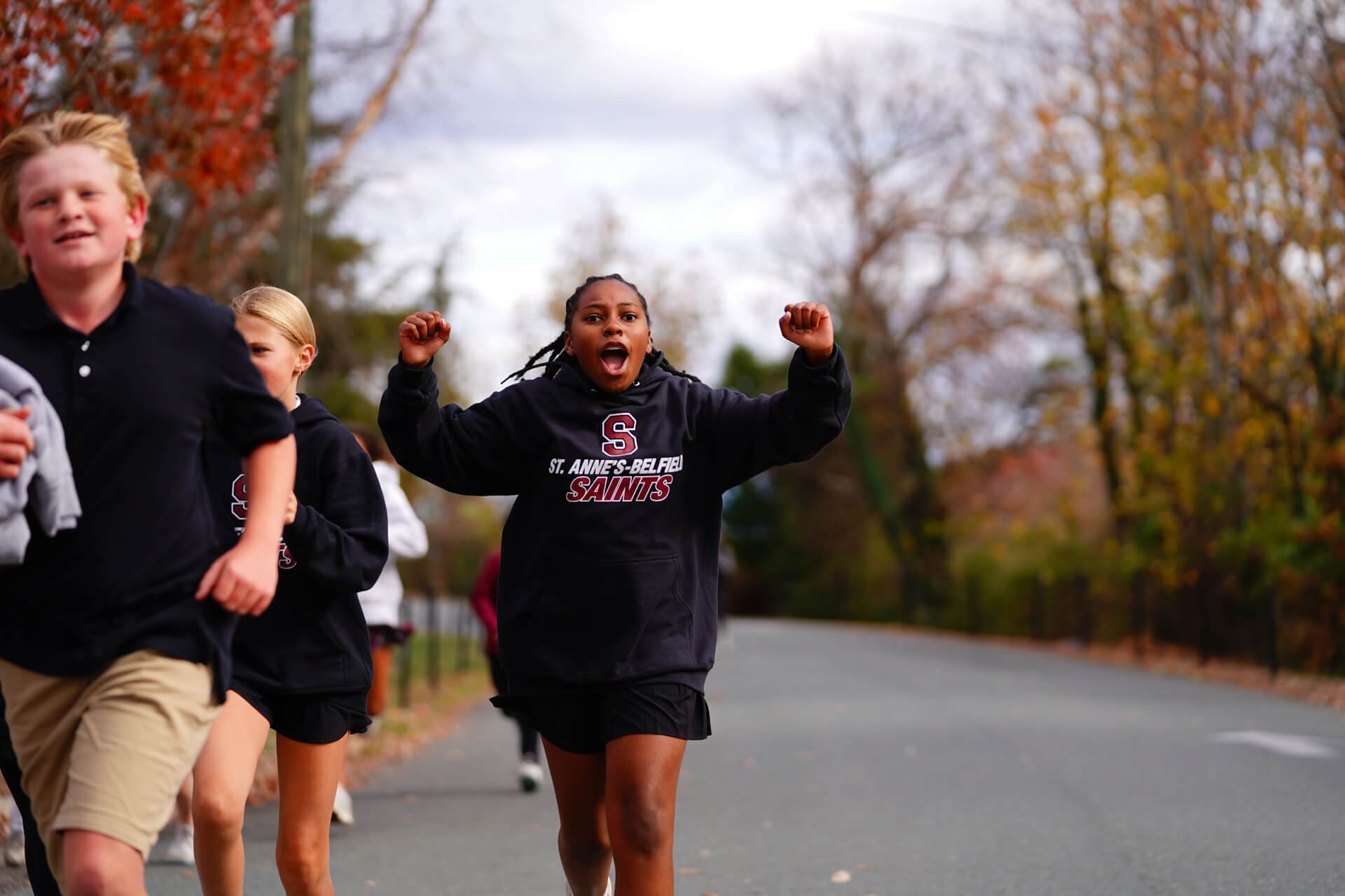 A group of young people runs outdoors on a road. One person in front, wearing a black Saints hoodie, raises their arms in excitement. Trees with autumn leaves line the background.