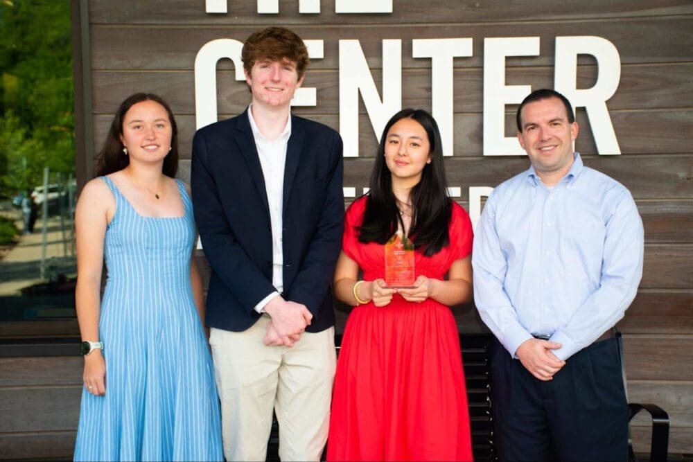Four people stand side by side smiling in front of a wooden wall with large white letters. One woman in a red dress holds an award. The others are dressed in formal attire, including a blue dress, a suit, and a light blue shirt.