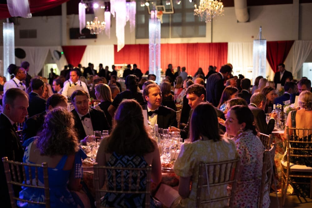 A large group of people in formal attire sit around tables set for dinner at an elegant indoor event, with chandeliers, red drapes, and warm lighting creating a festive atmosphere.