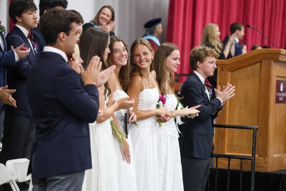 A group of young people in formal attire, including girls in white dresses holding flowers, stand and smile while others clap at an indoor ceremony with a podium and red curtains in the background.