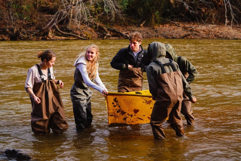 Five people wearing waders stand in a shallow river, likely as part of a school fieldwork project, holding a large yellow container. The group appears to be collecting samples, surrounded by trees and autumn foliage.