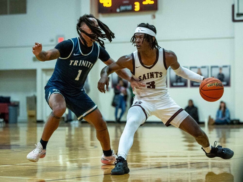 Two school basketball players in action on a court: one in a white Saints uniform dribbles the ball while being closely guarded by an opponent in a blue Trinity uniform. Spectators and a scoreboard are visible in the background.
