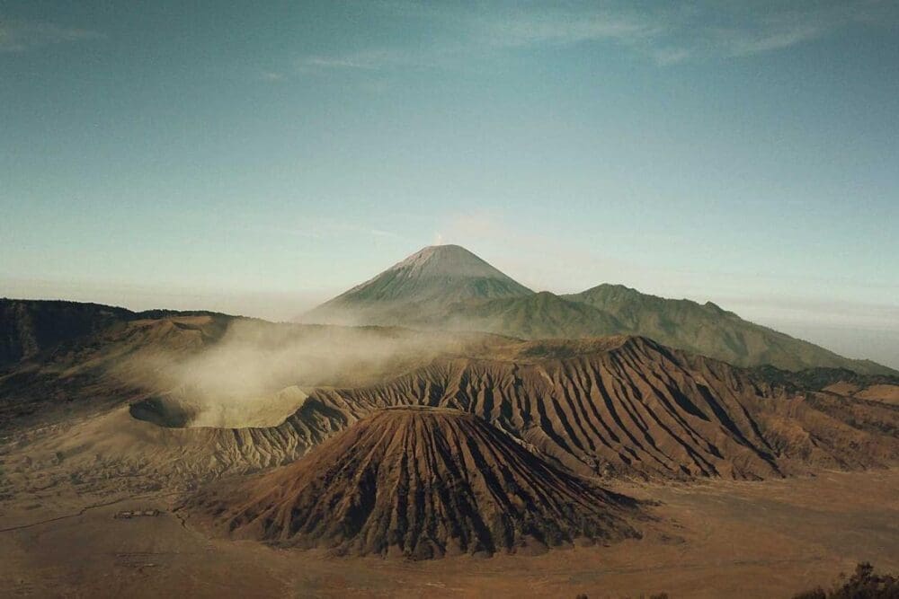 A wide view of a dramatic volcanic landscape with a steaming crater in the foreground, rugged ridges, and a tall volcano rising majestically in the background under a clear blue sky.