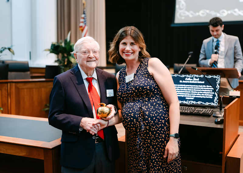 An older man in a suit holds a gold apple award and stands next to a smiling woman in a blue dress with white spots. They are in a formal setting with a man speaking at a podium and a projected screen in the background.