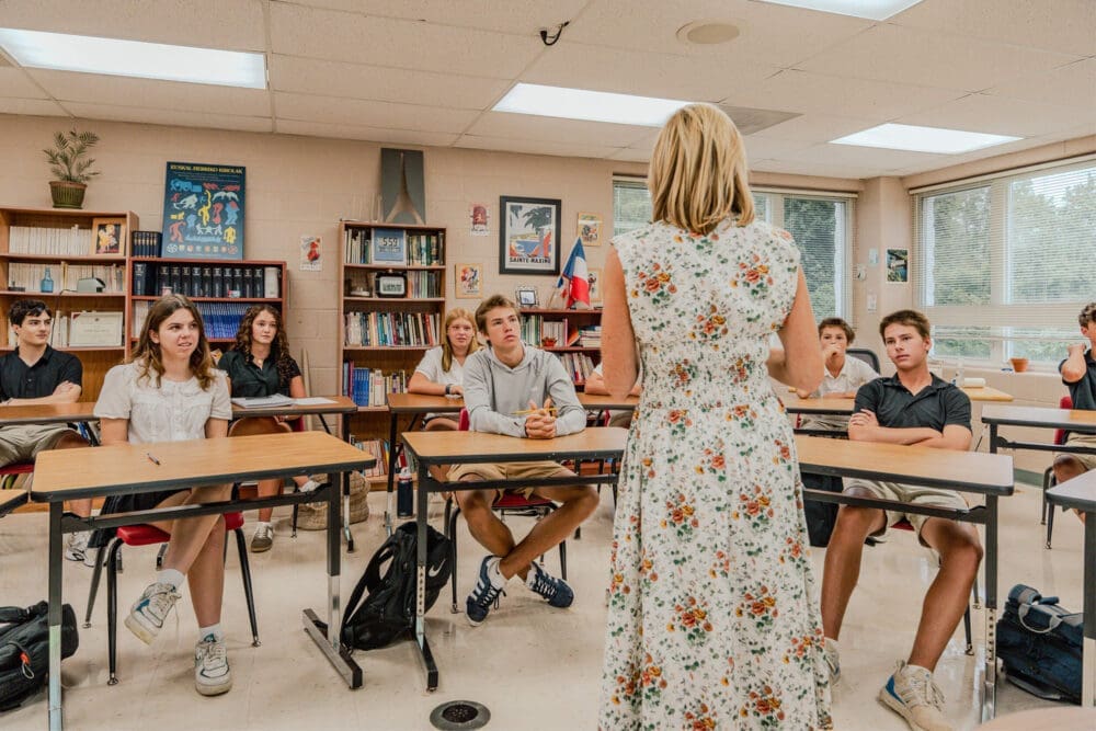 A teacher in a floral dress stands at the front of a classroom, speaking to attentive high school students seated at desks. Bookshelves and educational posters line the walls.