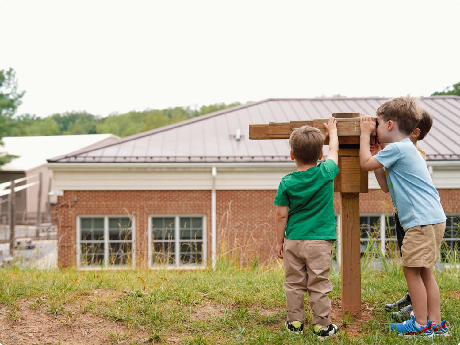 Three young boys stand outside on grass, looking through a wooden viewing structure. A large brick school building with a metal roof is in the background. The boys are facing away from the camera.